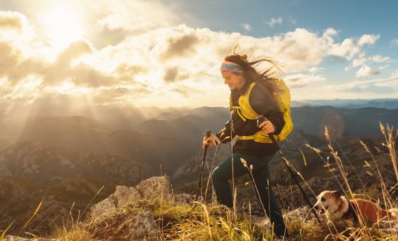 caucasian woman equipped with backpack, warm clothes and trekking poles hiking with her dog on a mountain at sunset. outdoor sports and adventures.